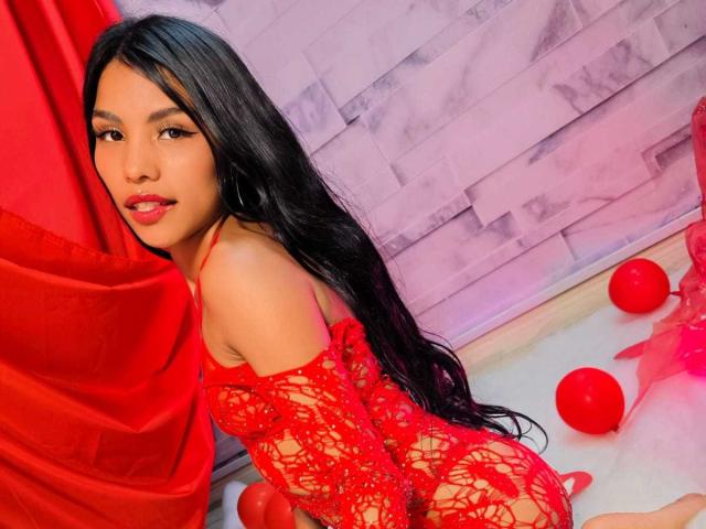 ClarisCampbell with long black hair, wearing a red lace lingerie set, sitting on the floor with red balls around her. Her red lips match the red fabric.