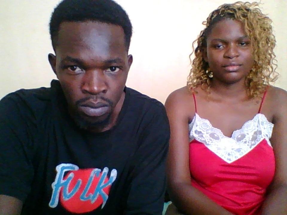 A photograph of a serious black man with short curly hair and a black T-shirt with "Funk" in blue and red letters, sitting next to a black woman with curly blonde hair wearing a red lace-trimmed spaghetti strap top. The man's dark skin contrasts with the woman's lighter skin. Both have intense, serious expressions. The woman's top has a white lace pattern. The man is on the left, the woman on the right.