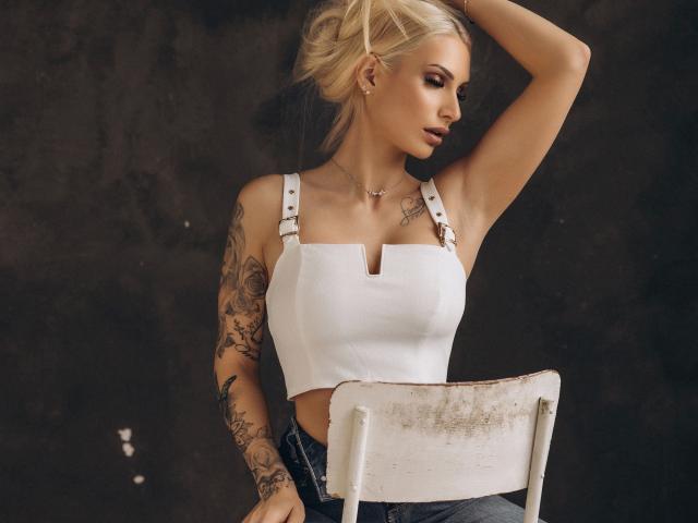 Lia-Juwel-hot with blonde hair, white crop top, tattoos on arm, holding white chair, wearing denim, hand in hair, looking to side, small tits, dark background.