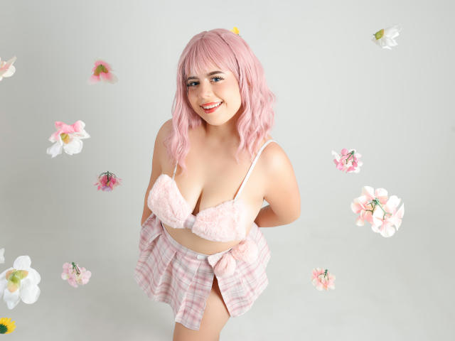 MelaniWare with pink hair, smiling, wearing pink lace bra, plaid skirt, and white underwear, hands behind back, surrounded by floating pink and white flowers.