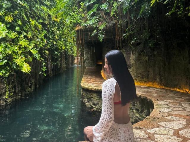 AngelaFoxxy sits on a stone path next to a clear, turquoise pool, her back to the viewer. She has long, straight black hair and wears a white, lace, long-sleeve top with a red bikini bottom. Lush green foliage and hanging vines surround the pool, with a dark, rocky cave entrance in the background. Light filters through the leaves, highlighting AngelaFoxxy's smiling profile and the calm water. The texture of the stone path contrasts with the smooth water and lace top.