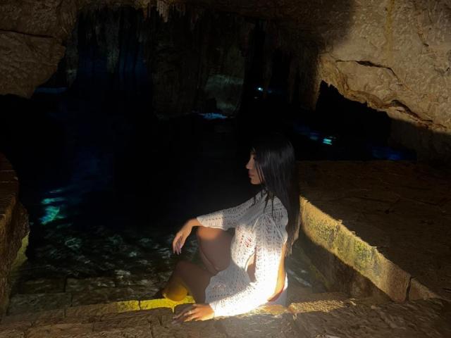 AngelaFoxxy sits in a dimly lit cave with rough, textured walls. She's in a white, crochet-patterned top and yellow socks, with long black hair. Her back is to the camera, highlighting her bare buttocks and legs. The cave's entrance casts a dark shadow, with a small, blue-lit pool of water in the background. The lighting emphasizes the texture of the cave walls and AngelaFoxxy's top.