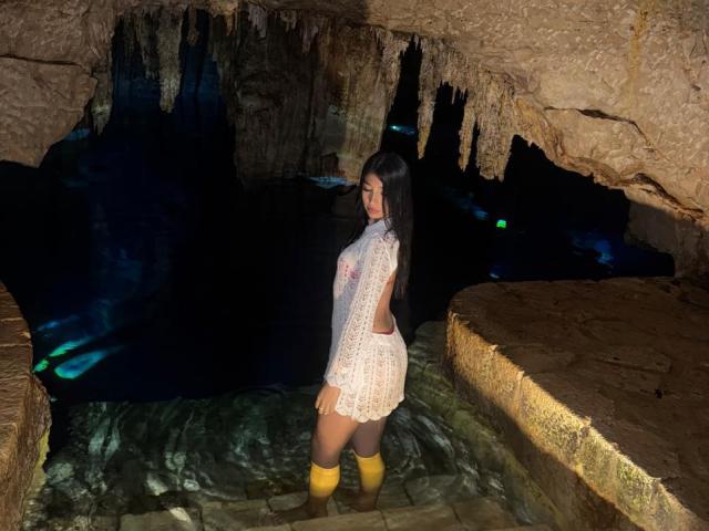 AngelaFoxxy with long black hair, wearing a sheer white lace dress, yellow knee-high socks, standing in a cave, water up to her thighs, showing her ass.
