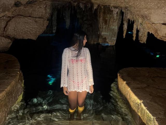 AngelaFoxxy stands in a dark cave, illuminated from behind. She's wearing a white, lace crochet top with pink floral patterns, and yellow knee-high boots. Her long black hair flows down her back. The rocky cave walls and ceiling have jagged, hanging icicles. The water she's standing in is clear, revealing the cave's stone floor. The contrast between the dark cave and her brightly lit top creates a striking visual.