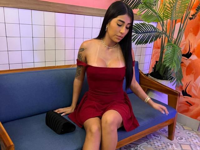 AngelaFoxxy with long black hair, red off-shoulder dress, small tattoo on right arm, sitting on blue couch, holding black purse, pink wall, white tiles, looking down, pearly necklace.