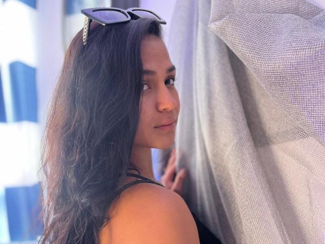 RouxiMori with long dark hair, wearing black tank top, black sunglasses on head, looking back with slight smile, hand on sheer curtain, soft natural light.