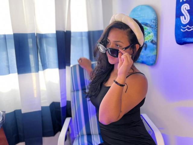RouxiMori with black dress, white hat, and sunglasses, adjusting black frames on nose, sitting on striped chair, blue and white curtains, and blue wall art in background.