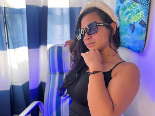 RouxiMori with dark hair, black tank top, and white beret, wearing black Ray-Ban glasses, touching her chin, showing her tits, and blue-striped chair in background.