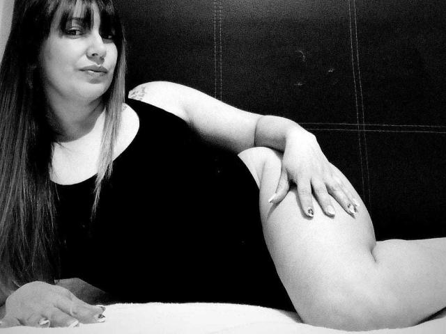 Black-and-white photo of RoxanneBlair with long hair, nose ring, and black one-piece, lying on bed, hand on thigh, displaying her ass, smiling slightly.