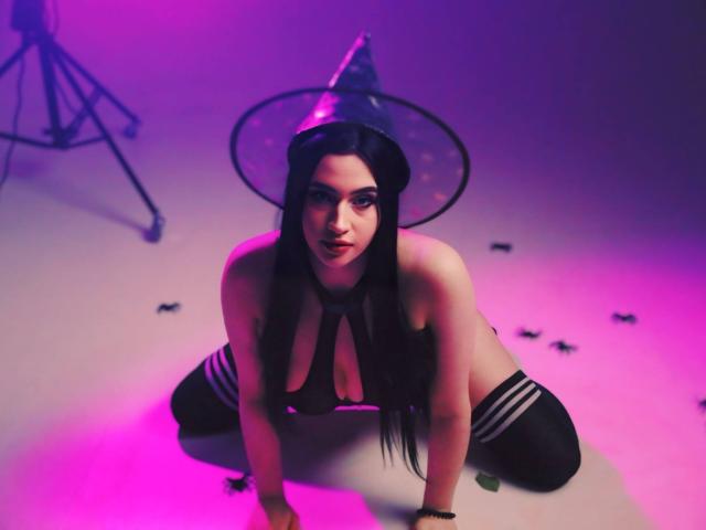 MaryLori, on knees, wearing a witch hat, black choker, and thigh-highs, with visible boobs, under purple and pink lighting, looking up, hands on floor, legs apart.