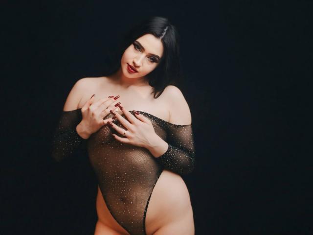 MaryLori wearing a sheer black mesh bodysuit with sparkles, covering her tits with her hands, standing against a dark background, looking down with red lipstick and black hair.
