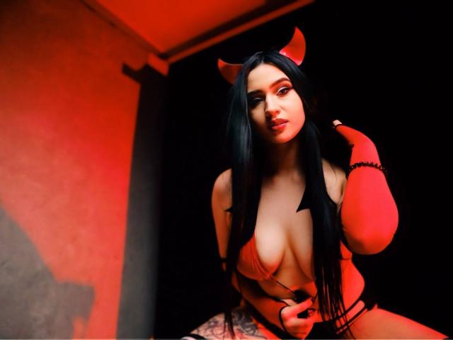 MaryLori with black hair, red devil horns, and a red bra, showing her tits, wearing red arm warmers, and a black thong, in a red-lit room.
