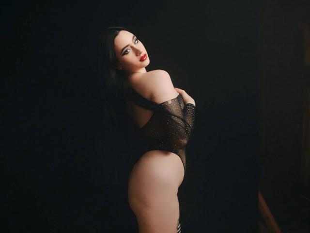 MaryLori with black hair, red lipstick, wearing a sheer black bodysuit, exposing her round ass and curvy hips, standing against a dark background.