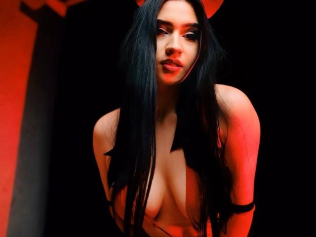 MaryLori with long black hair, red lighting, and a black ribbon over her tits, leaning forward, looking at the camera with a slight smile.