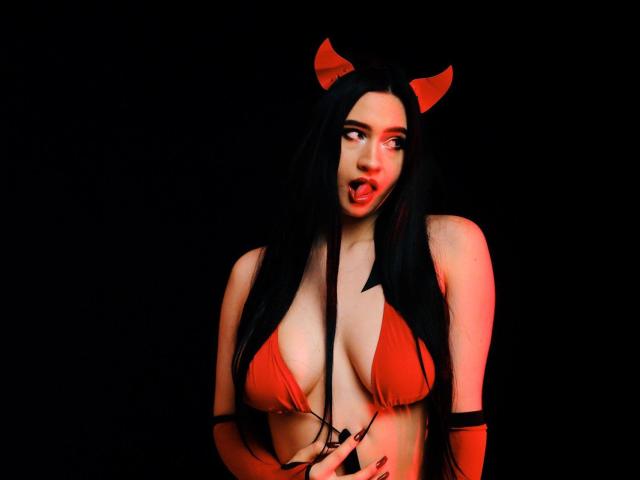 MaryLori with long black hair, red devil horns, red bikini top showing big tits, red fingerless gloves, black suspenders, and red lipstick, against a black background.