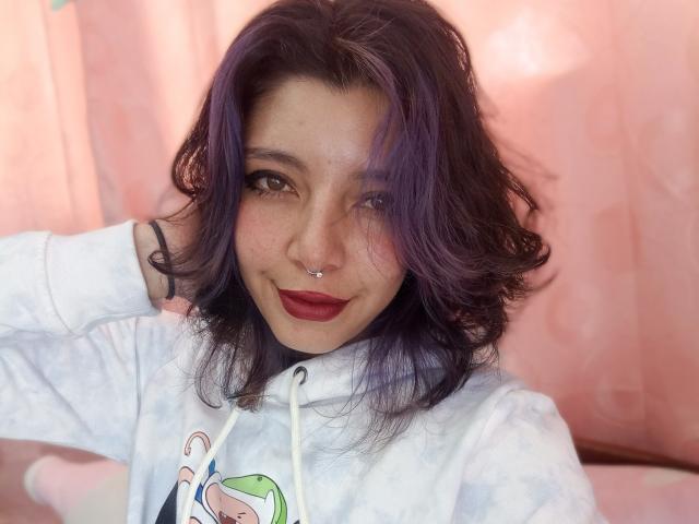 DarckVelvet has purple hair, brown eyes, a nose ring, and red lipstick, wearing a white hoodie with a cartoon design, hand in hair, smiling.