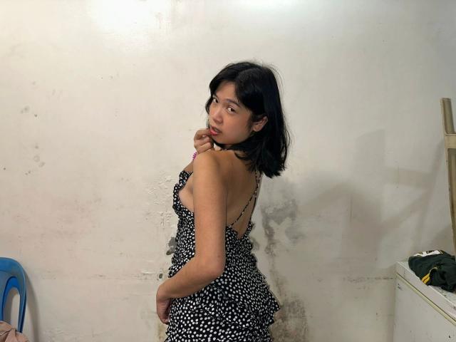 AsianSlut-hot with short black hair, wearing a black and white polka dot dress, looking over shoulder, biting finger, standing against a white, slightly dirty wall.