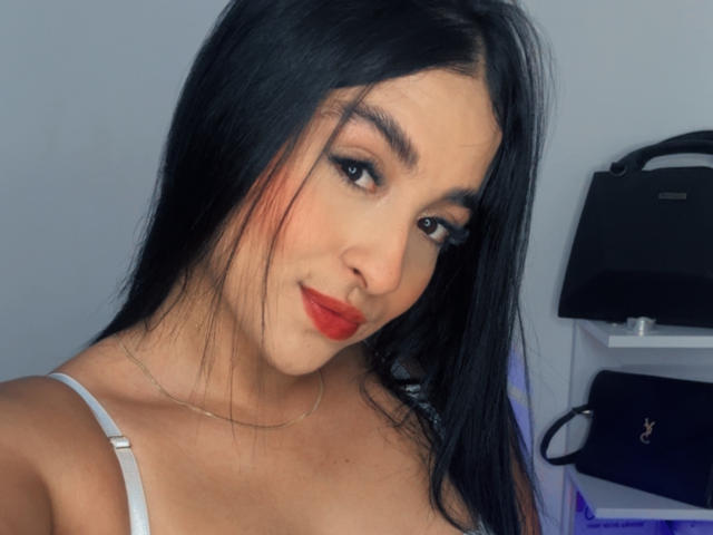 EmmaWaltter with black hair, red lipstick, white strap top, looking at camera, slight smile, medium tits, light skin, subtle makeup, in a selfie with a black handbag in background.