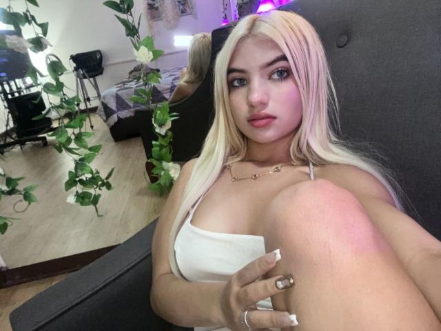 SabrinaColle, con i capelli biondi, gli occhi azzurri e le labbra rosa, indossa una maglietta bianca, è seduta con le ginocchia sollevate, mostrando il seno, su un divano grigio, con una pianta sullo sfondo..
