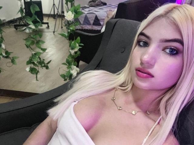 SabrinaColle with blonde hair, pink lips, blue eyes, white tank top showing her tits, necklace, sitting in gray chair, looking at camera, wooden floor, green vines in background.