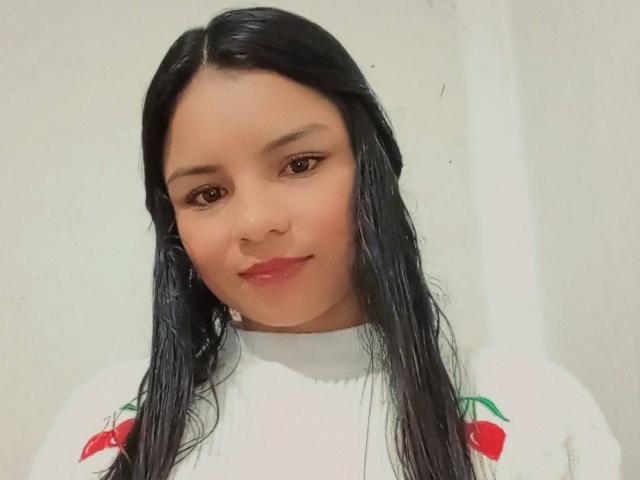 CrystalStones has wet black hair, brown eyes, and red lipstick, wearing a white sweater with red cherry designs. She's smiling softly.