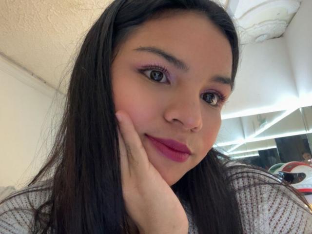 CrystalStones has long black hair, pink eyeliner, and pink lipstick, with her hand gently touching her cheek. She has a cute smile and is wearing a white knitted top.
