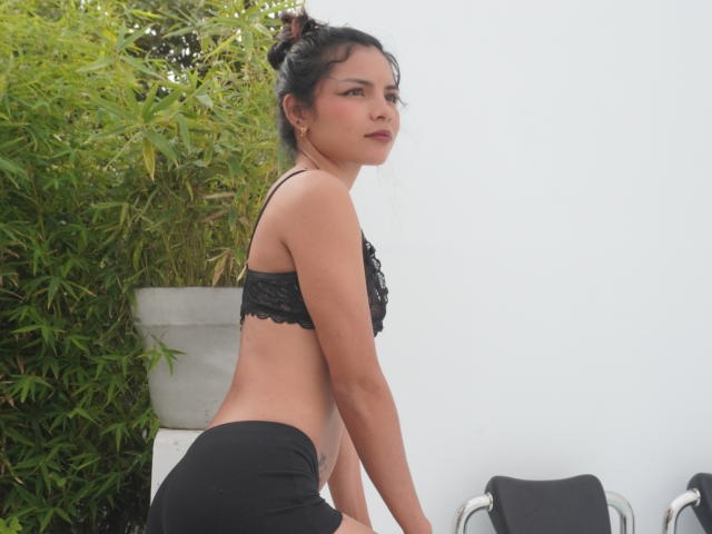CrystalStones with dark hair in a bun, black lace bra, and black shorts, leaning forward, showing her ass, and looking over her shoulder. Her breasts are small.