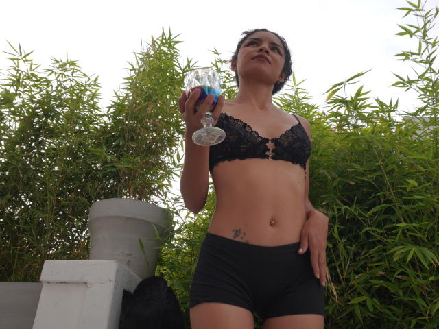 CrystallStones with small tits, black lace bra, black shorts, holding a glass of blue drink, looking up, tattoo on right hip, bamboo in background, standing outdoors, daytime.