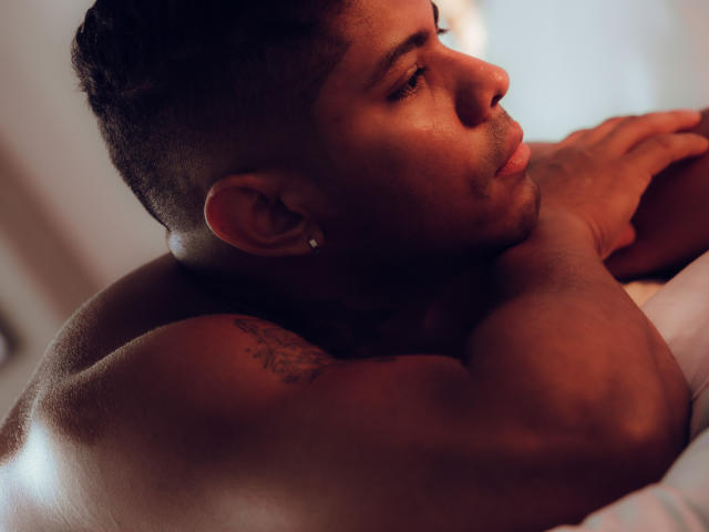 LeonHrz, shirtless, muscular, brown skin, short dark hair, small ear tattoo, side profile, resting arms, soft light, focused expression, bed in background, subtle shadows.