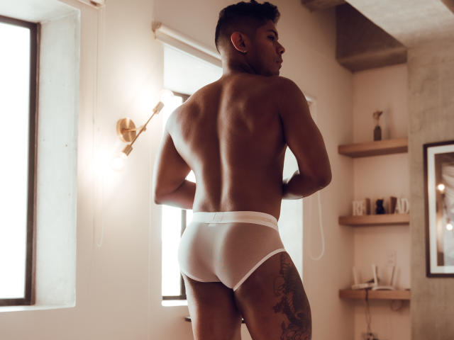 LeonHrz, muscular, dark-skinned, with a tattoo on his right thigh, wearing white, tight, bulging, butt-focused underwear, standing with his back to a window, looking over his shoulder.