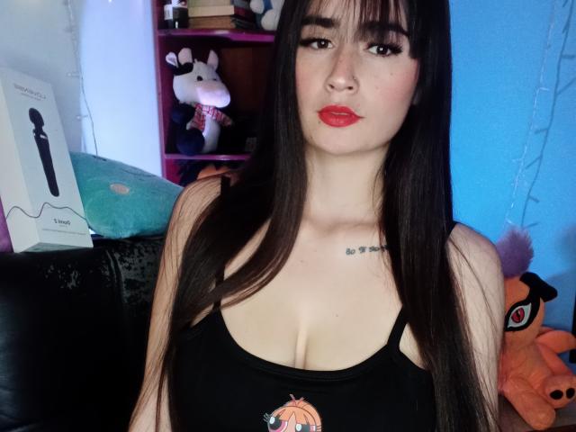 EllieBonneth has long dark brown hair, red lipstick, and a black tank top showing small tits with a pumpkin design. She's pouting with a tattoo on her chest.