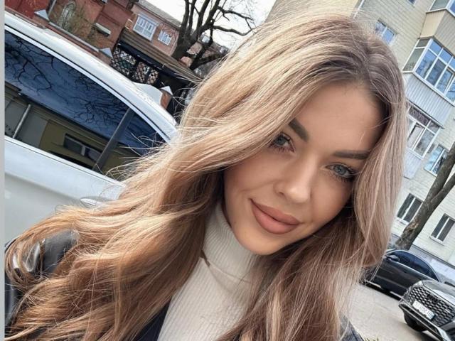 LizaSweety with long, wavy blonde hair, blue eyes, and full pink lips, wearing a white turtleneck, standing next to a white car. She's smiling softly.