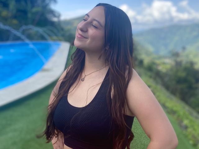 RosalieDupont with long brown hair, smiling, wearing a black sports bra, showing her tits, by a blue pool, green grass, and mountains in the background.