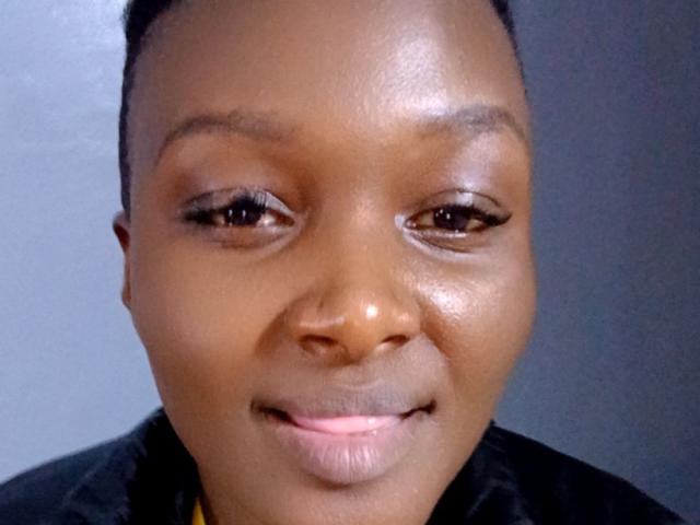 Close-up of SexysChatz with a medium-dark brown complexion and short, neat hair. SexysChatz has subtle, natural makeup with defined eyebrows, small eyelashes, and a slight smile showing pink lips. SexysChatz wears a black shirt with a yellow collar visible at the bottom. The image has a slight reflection on the face, emphasizing smooth skin texture. The overall composition is centered with a neutral, slightly grey background. The lighting is even, highlighting SexysChatz's facial features and skin tone.