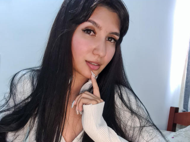 NaylaGrey, com cabelos pretos longos, pele clara e olhos castanhos, tocando o dedo nos lábios, vestindo uma camiseta branca texturizada, exibindo seus seios de forma discreta..