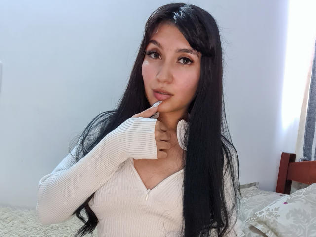 NaylaGrey with long black hair, wearing a white ribbed long-sleeve top, biting her finger, showing her tits, sitting on a bed with a white wall behind.
