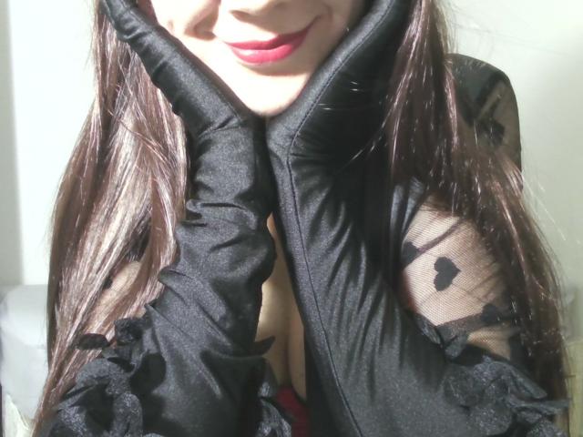 Close-up of AromaLove's face and chest. She has long, dark brown hair, and her lips are painted red. She wears a black, sheer, and ruffled top with floral patterns on the sleeves. Her hands are near her face, with black, textured sleeves covering them. The top's fabric is slightly wrinkled, and a hint of red is visible at the bottom. Bright light casts shadows, emphasizing her hair and top's texture.