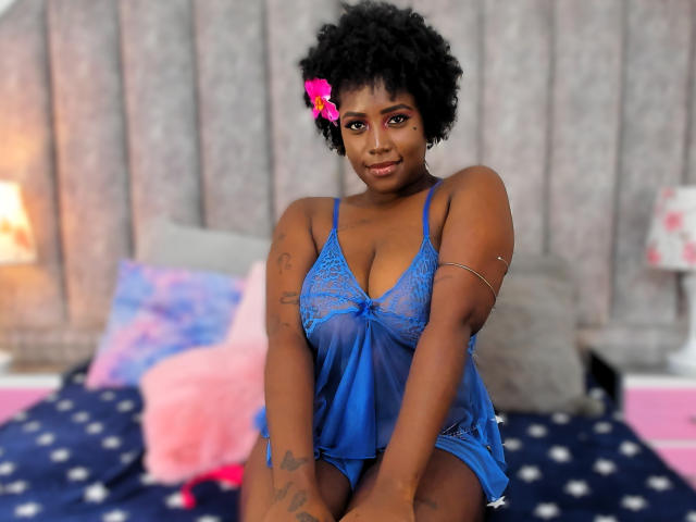 EimmyCampbell with dark skin and curly black hair, wearing a blue lace babydoll, has small tits and tattoos, sitting on a bed with pink and blue pillows.