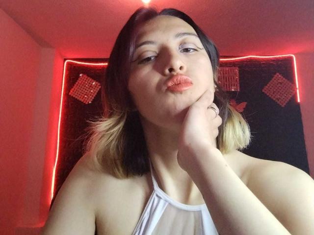 SylvieCute with pouty lips, wearing a white halter top, black and blonde hair, hand on cheek, red lighting, and a ring on her finger.