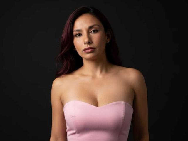 RoxaneMoore with medium tits in a pink strapless dress, dark purple hair, and a serious expression, standing against a black background.