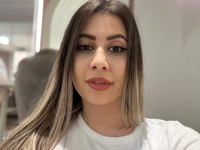 Close-up of DiamondHottieX. She has straight, long brown hair with subtle highlights, dark eyebrows, and thick, black eyeliner. Her lips are painted a soft red. She wears a white shirt. The photo is well-lit with a slight shadow on her left cheek. The background is blurred, focusing attention on her face. The image is sharp with a clear, centered composition.