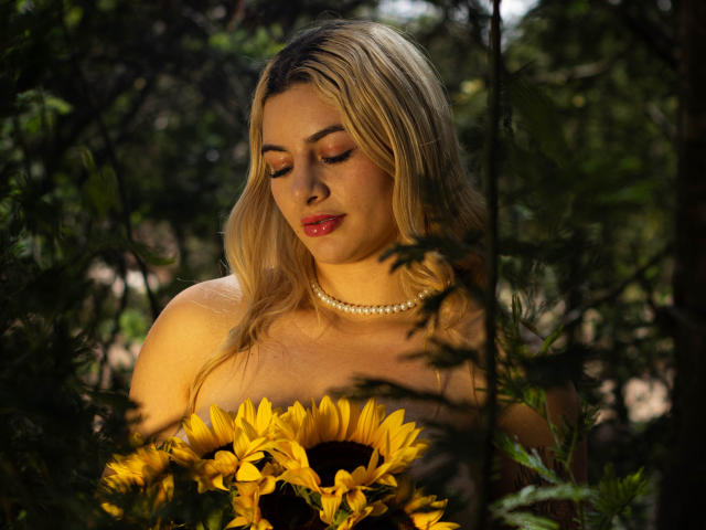 AtheneaRozzi with blonde hair, red lipstick, and pearl necklace, holding sunflowers, looking down, surrounded by greenery, in a dimly lit, natural setting.