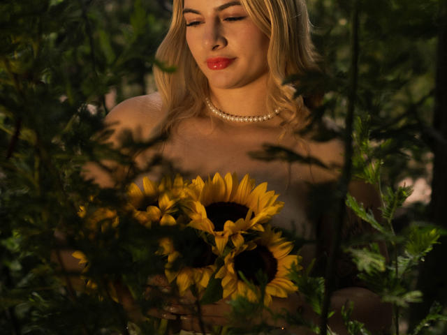 AtheneaRozzi with blonde hair, red lipstick, and pearl necklace, holding sunflowers, standing in greenery, soft light, gentle smile, showing bare shoulders, looking down at sunflowers.