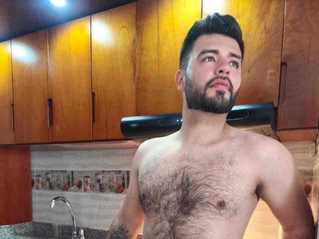 Salvathore, shirtless with a hairy chest, dark beard, and short black hair, stands in a kitchen with a black range hood, looking to the side.