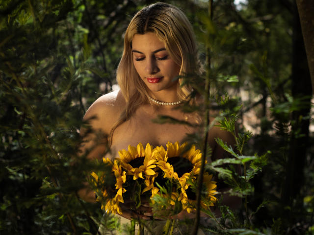AtheneaRozzi with blonde hair, holding sunflowers, wearing a pearl necklace, looking down, standing in a green forest, sunlight on her face, soft focus, natural light.