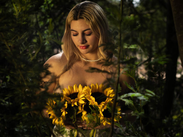 AtheneaRozzi with blonde hair, pearl necklace, and red lipstick, holding a sunflower bouquet, her bare shoulders and tits visible, standing in a green, forest-like setting.