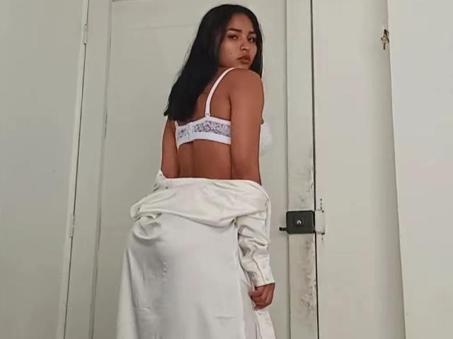 A photograph of BellaChaudAmour with medium brown skin and long black hair, looking over her shoulder. She's wearing a white bra with "Hollister" text and a white shirt, which is partially off her shoulders, revealing her back. The bra straps are visible on her shoulders. The white door in the background is slightly worn. Her expression is neutral, and she's centered in the frame, with the door to her right. The image is clear and well-lit.