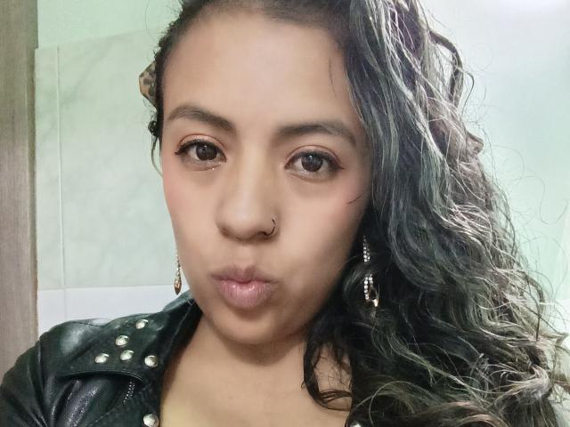 MeganFerrerr with wavy dark hair, nose ring, and pouty lips, wearing a black leather jacket with studs, and dangling earrings, in a selfie.