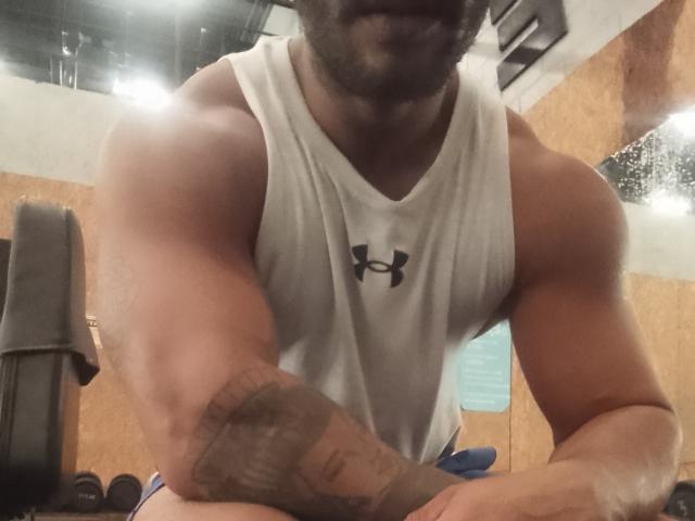 Kanekki, muscular, tattooed arm, white Under Armour tank top, arms crossed, black beard, partial face visible, indoor gym setting, focused on Kanekki's upper body.