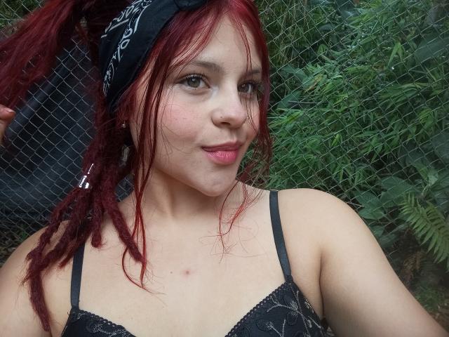 SophieHill with red hair, black bandana, and black lace bra, pouting with pink lips, slight freckles, and small red marks on chest, standing in front of a chain-link fence.